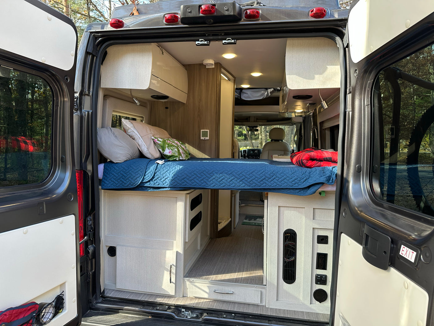 Spacious RV rental interior featuring a cozy bed, cabinetry, and controls, viewed from the open rear doors. Ideal for adventures.