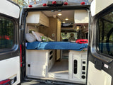 Spacious RV rental interior featuring a cozy bed, cabinetry, and controls, viewed from the open rear doors. Ideal for adventures.