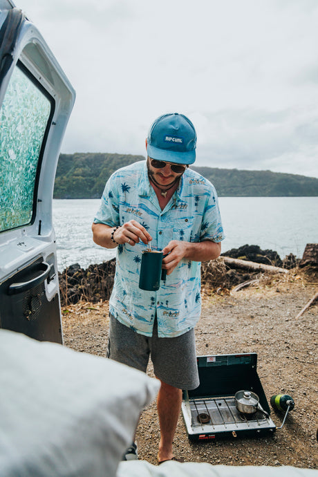 A man in a Rip Curl hat and Hawaiian shirt stirs coffee from an rv rental, cooking on a stove by the water and lush hills.