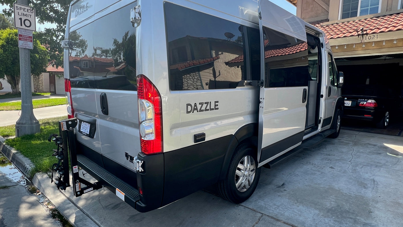 Silver Dazzle camper van RV rental with open side door, parked on a driveway. Features a rear bike rack, near street signs.