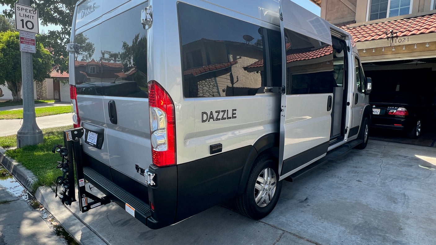 Silver Dazzle camper van RV rental with open side door, parked on a driveway. Features a rear bike rack, near street signs.