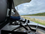 An RV rental's compact kitchen with a stove and sink offers scenic views of a lake and forested hills.