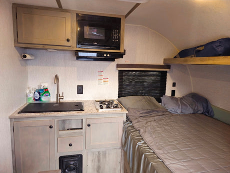 RV rental interior with compact kitchen, sink, stove, microwave, bed, and an overhead bunk.