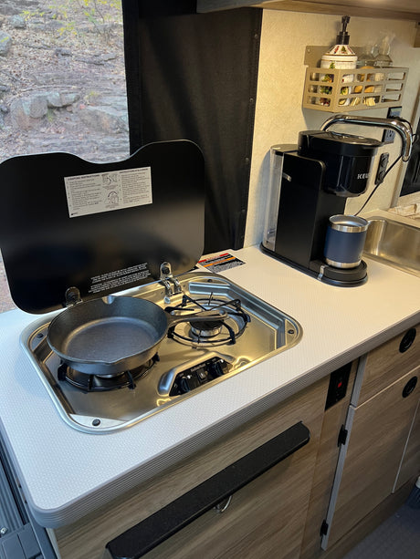 Inside an RV rental: a compact kitchen with a built-in stove, cast iron pan, Keurig coffee maker, and sink.