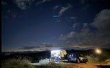 Night camping with an RV rental and SUV, lit under a starry sky. Green and blue lights are visible among the clouds.
