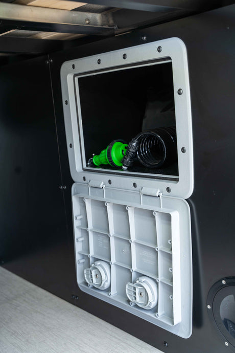 Inside an RV rental: open exterior utility access hatch revealing a coiled hose with green nozzle & two connection points.
