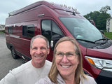 A happy couple smiles, taking a selfie in front of their maroon RV rental van, ready for travels.