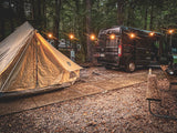 Evening **RV rental** camping setup: a cozy bell tent and black camper van illuminated by string lights in a wooded campsite.