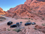 Remote red rock desert camping area with a white camper van (rv rental), multiple trucks, and tents along a dirt road.