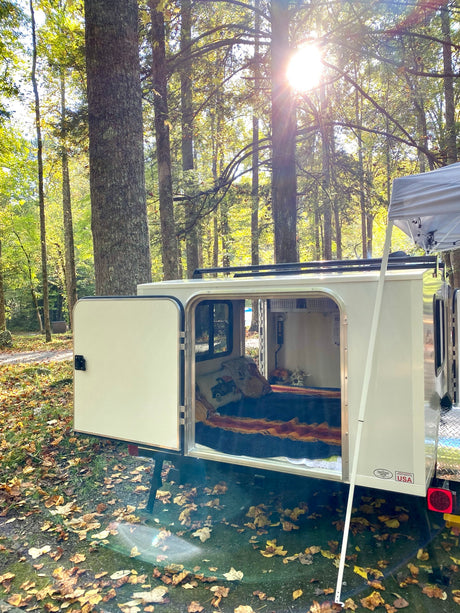 Experience nature with this rv rental! A cozy teardrop camper with its door open reveals a bed, nestled in a sunny fall forest.