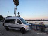 Silver Sprinter RV rental by waterfront at sunset. Palm trees frame this adventure-ready camper van.