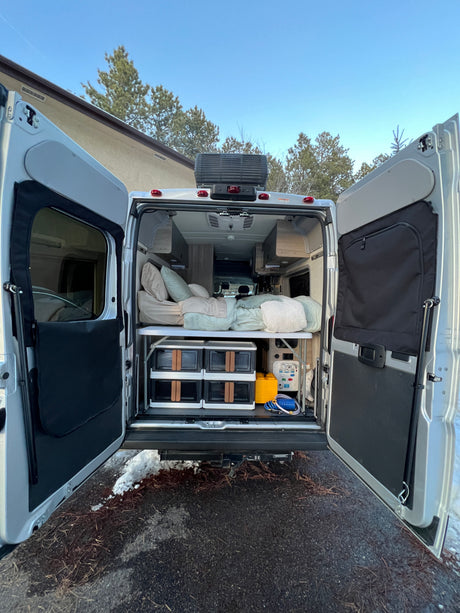 Open rear of an RV rental showing a cozy bed, organized storage, and utility panel for comfortable adventures.