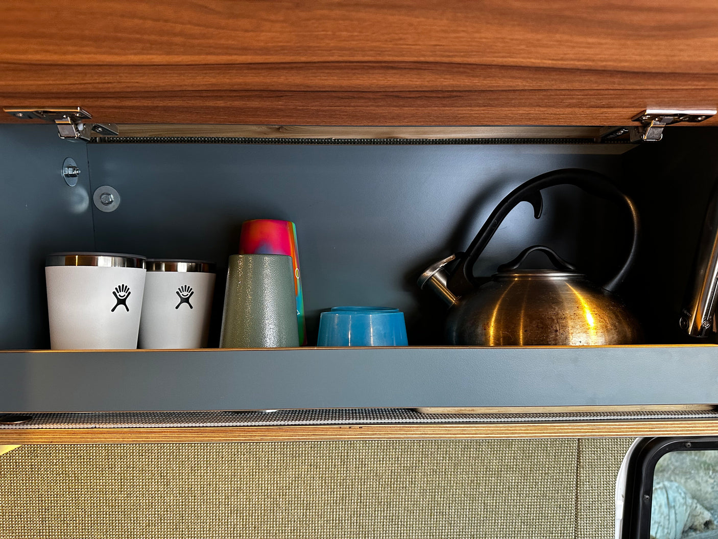 Inside an RV rental cabinet: Hydro Flasks, colorful cups, and a kettle neatly stored, ready for travel.
