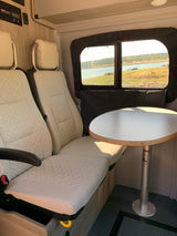 Cozy interior of an RV rental with cream seats, a round table, and a scenic view of a lake through the window, ideal for travelers.