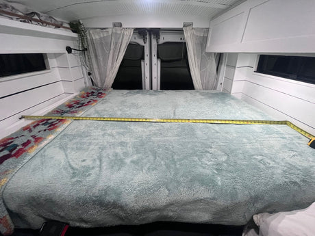 Inside an RV rental camper van: a cozy, light blue bed with a tape measure showing its width, nestled between white walls and curtains.