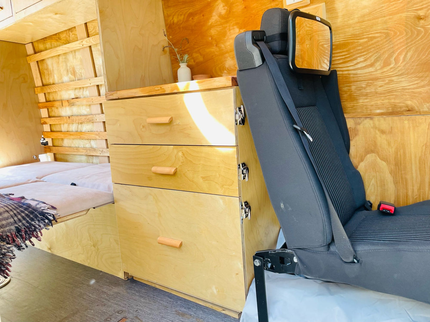 RV rental: Bright, wooden van interior with built-in storage drawers, a cozy bed, and a vehicle seat with mirror. — photo 20