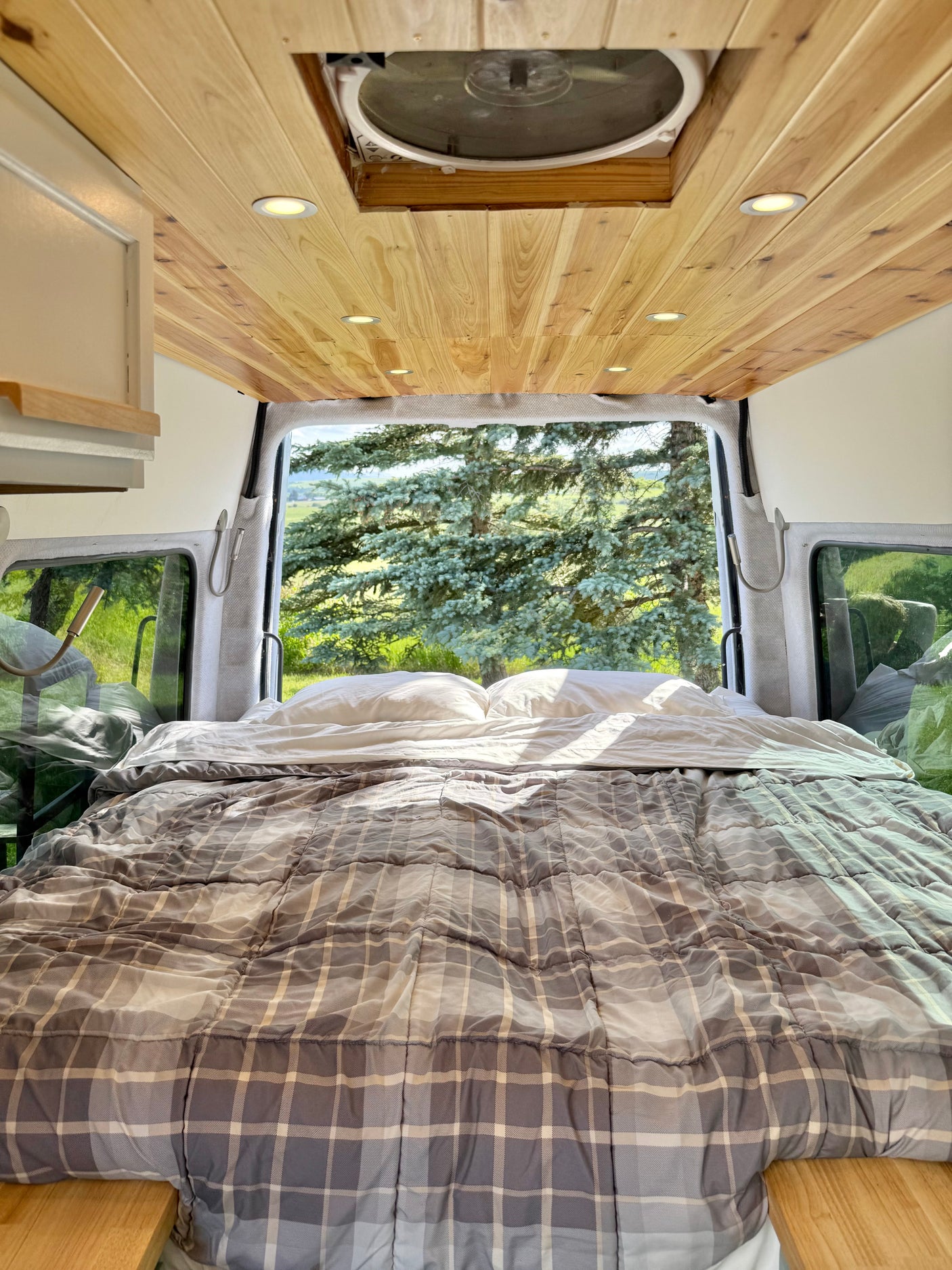 Cozy RV rental bedroom in a camper van, featuring a plaid bed, wooden ceiling, and an expansive nature view from the open back.