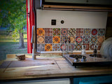 Inside a modern RV rental kitchen: wooden countertop, stainless sink, colorful tile backsplash, and cooktop, with a view of trees.