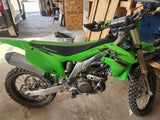 Bright green Kawasaki KX250X dirt bike, mud-splattered & ready for trails, parked inside near an RV rental.