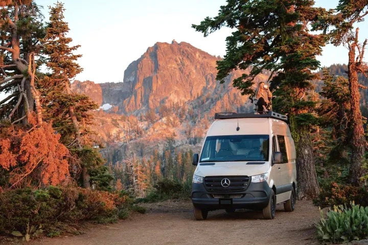 RV rental adventure: Tan camper van parked with person on roof enjoying stunning mountain views at golden hour.|RV rental: Cozy camper van bedroom with a stunning mountain view from the back — photo 1