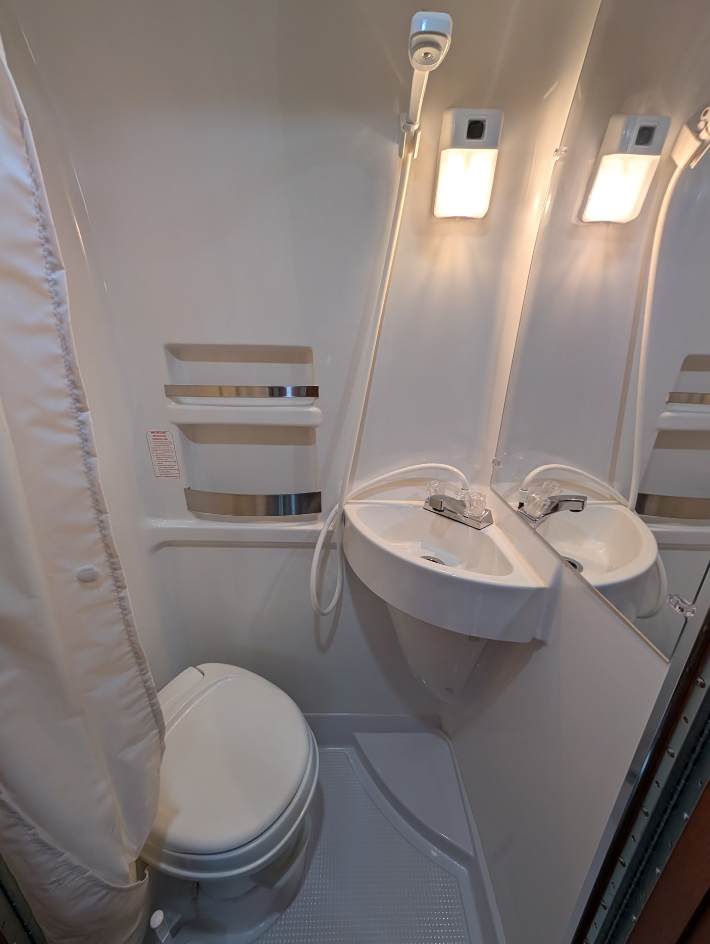 This RV rental wet bath includes a toilet, corner sink with shower hose, mirror, and bright lights, ideal for compact travel.