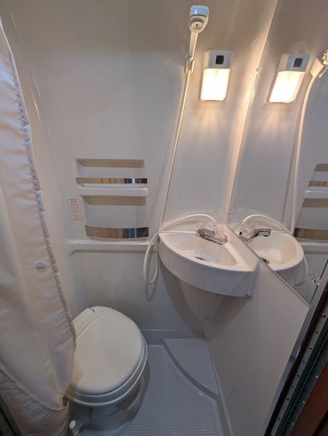 This RV rental wet bath includes a toilet, corner sink with shower hose, mirror, and bright lights, ideal for compact travel.