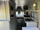 RV rental interior: compact kitchen, bed, & storage in a modern camper van, featuring gray fabric walls and a clean design.