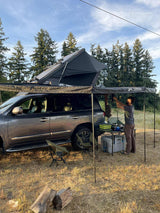Man sets up an outdoor kitchen from an SUV with a rooftop tent & awning – a compact RV rental-style camp.