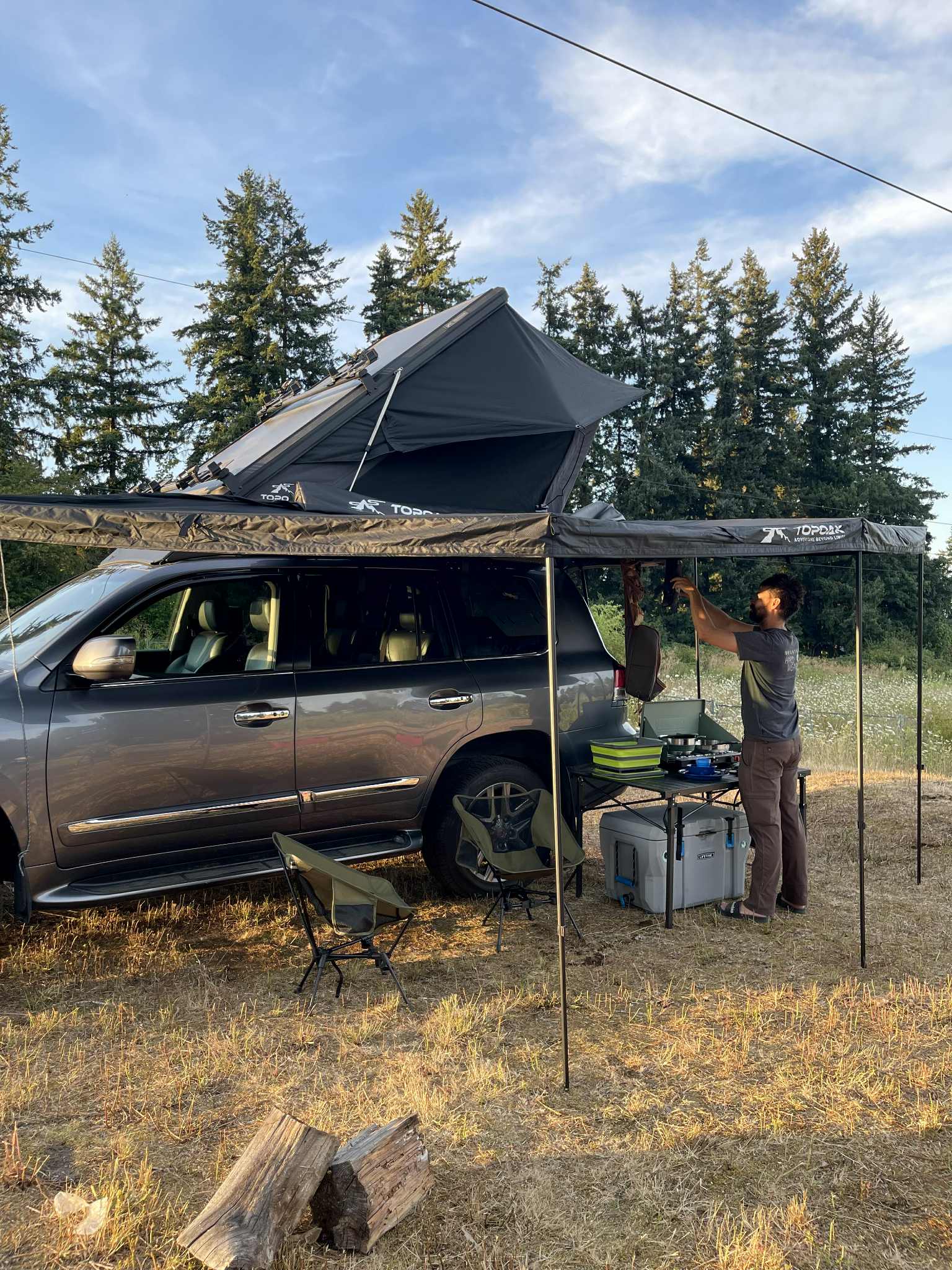 Man sets up an outdoor kitchen from an SUV with a rooftop tent & awning – a compact RV rental-style camp.