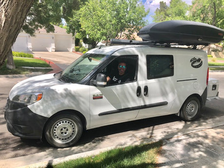 Explore with this white camper van, ideal for RV rental adventures, featuring a roof cargo box & smiling driver.