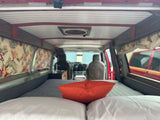 Cozy RV rental interior shows a bed with an orange pillow, floral curtains, and custom wood details.