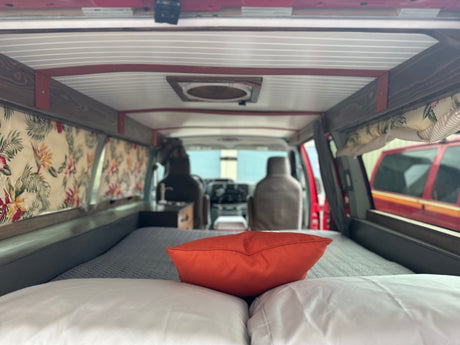 Cozy RV rental interior shows a bed with an orange pillow, floral curtains, and custom wood details.