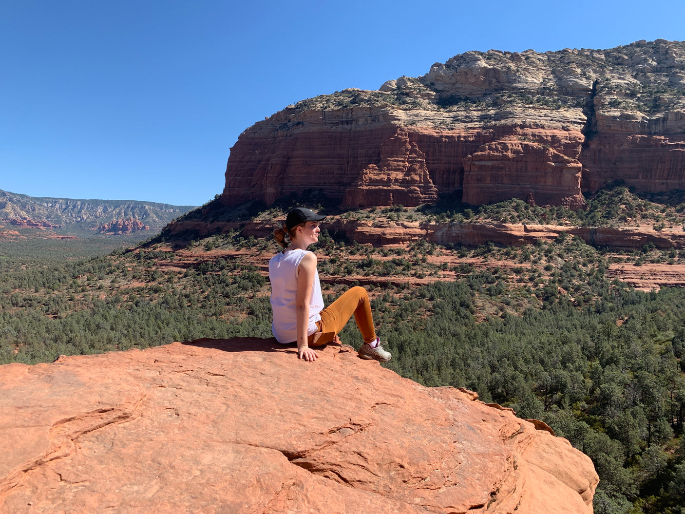 Woman sits on red rock, admiring a vast, forested red rock landscape under blue skies. Ideal stop for an RV rental trip.