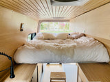 Interior of an RV rental camper van showing a large bed with white bedding, light wood walls, and a view outside.