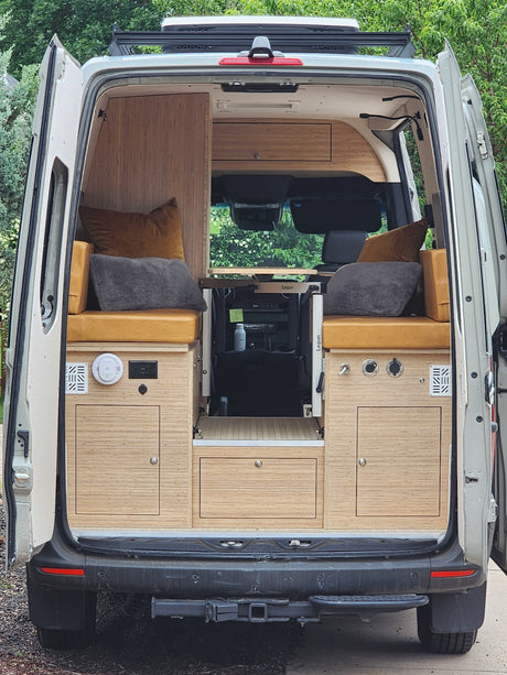 Rear view of an open RV rental camper van's interior. Shows custom bamboo cabinetry, bench seating, a flexible table, and integrated utilities.