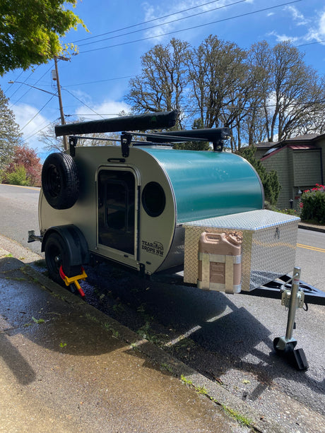 A green and grey teardrop **rv rental**, secured with a wheel lock, sits curbside ready for adventure.