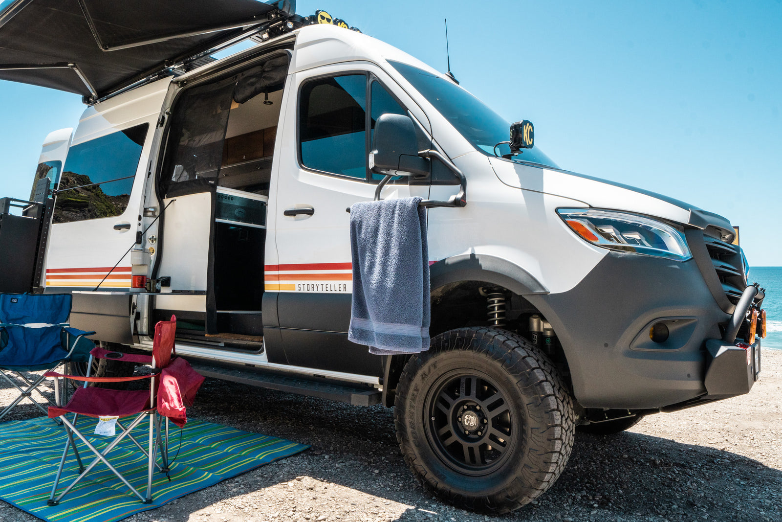 White Storyteller RV rental with awning extended, side door open to camping chairs and mat, parked at a sunny beach.