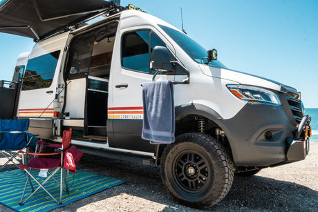 White Storyteller RV rental with awning extended, side door open to camping chairs and mat, parked at a sunny beach.