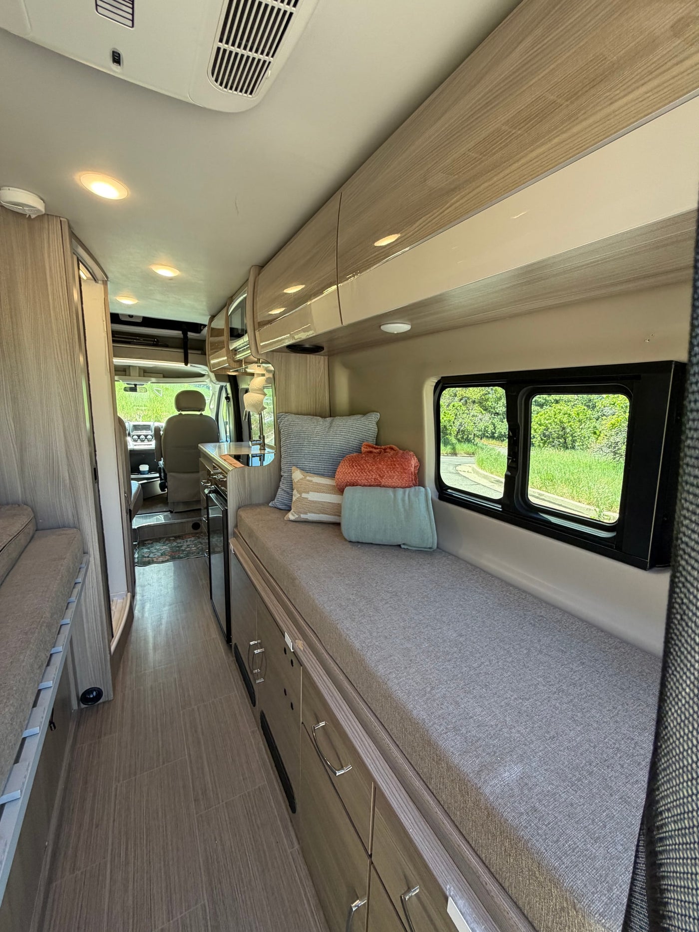Bright RV rental interior: cozy bench with pillows, kitchen area, wood-like floor, overhead storage, and driver's cab view. — photo 10