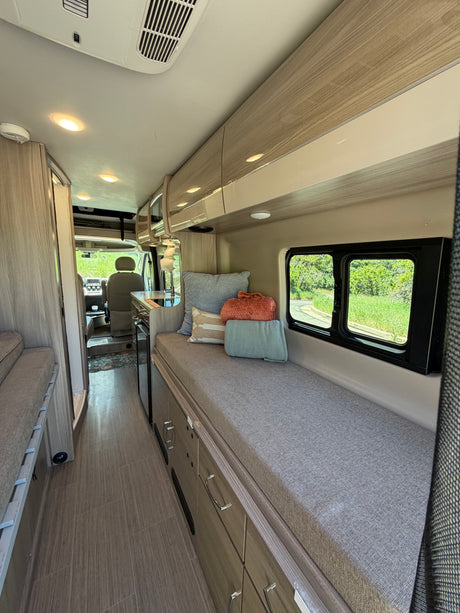Bright RV rental interior: cozy bench with pillows, kitchen area, wood-like floor, overhead storage, and driver's cab view.