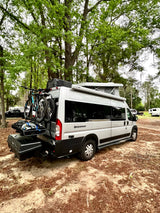 A silver "Sequence" RV rental with a pop-top, bikes, and cargo is parked in a wooded campground setting.