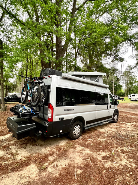 A silver "Sequence" RV rental with a pop-top, bikes, and cargo is parked in a wooded campground setting.
