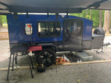 Blue camper trailer with an extended awning, propane tank, and outdoor cooking setup at a gravel campsite. Great for rv rental adventures.