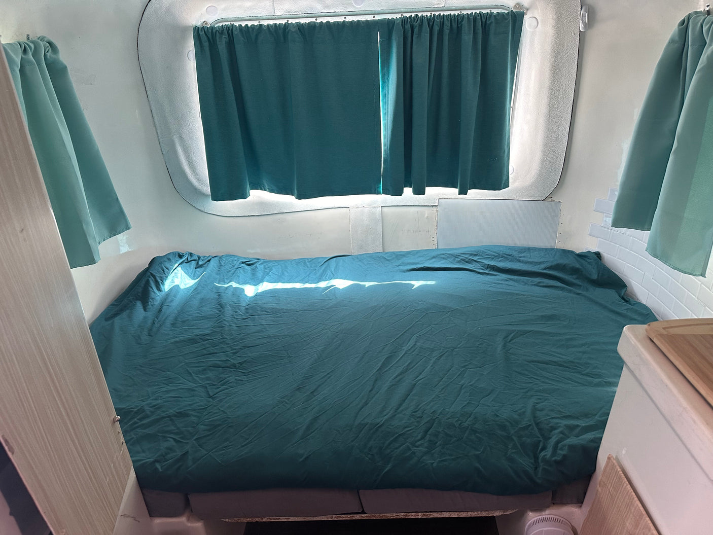 Comfortable sleeping area inside an RV rental, featuring a bed with teal covers and matching curtains. Sunlight illuminates the space.