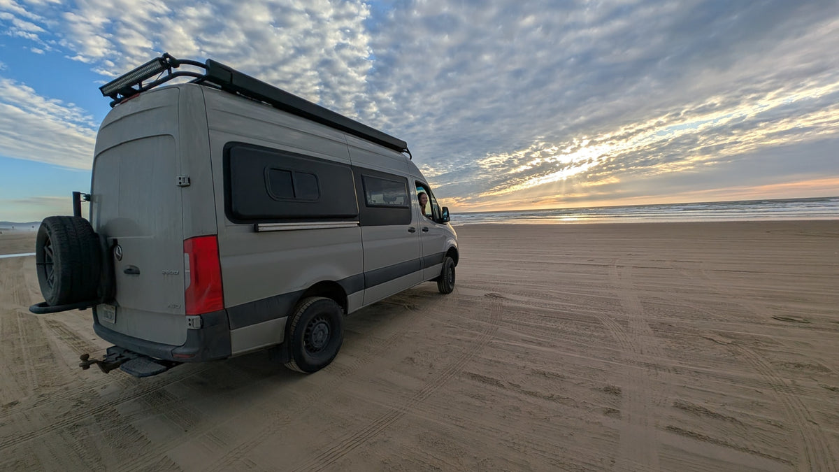 This gray RV rental campervan awaits sunset adventures on a wide sandy beach, a person visible inside, ocean waves rolling. — photo 4