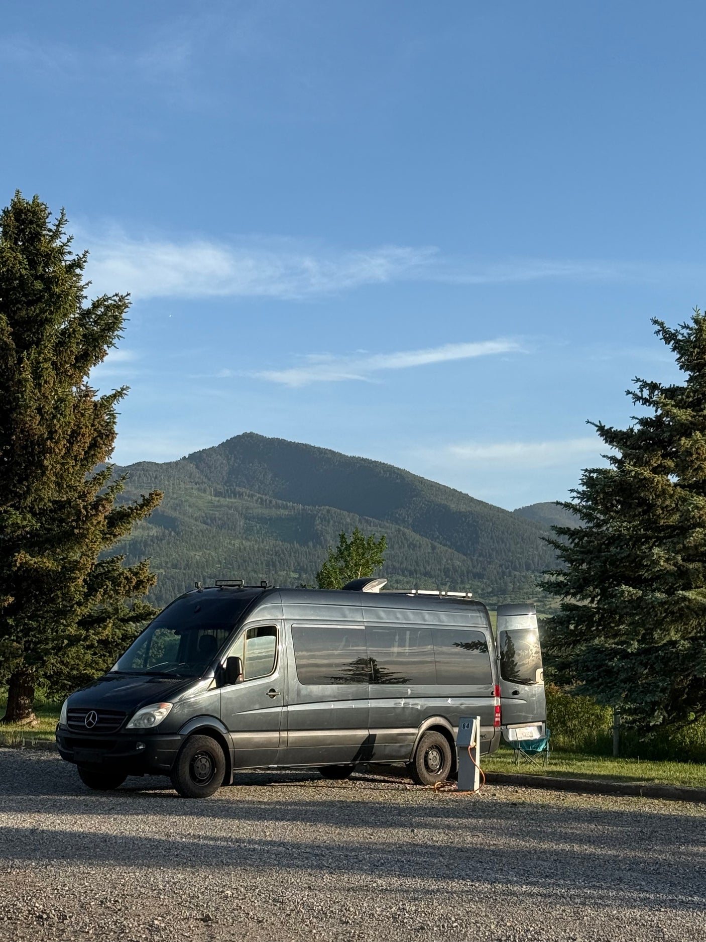 Gray Sprinter van, likely an RV rental, parked on gravel with pine trees and mountains under a blue sky.