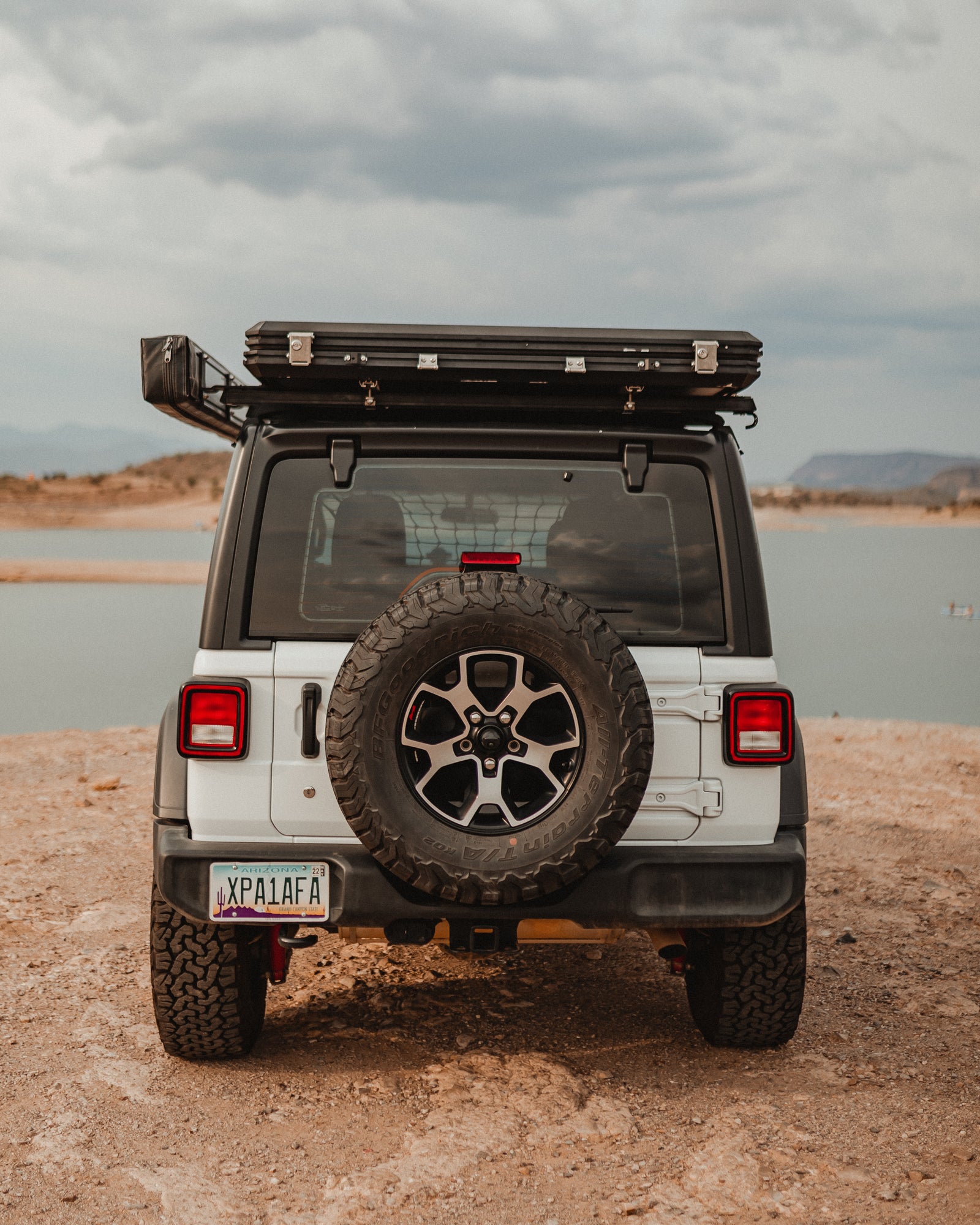 A white Jeep Wrangler with a rooftop tent, ideal for an RV rental adventure, is parked by a scenic lake under cloudy skies.