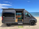 This gray RV rental is perfectly parked on a sandy beach, its open side door revealing a cozy custom interior.