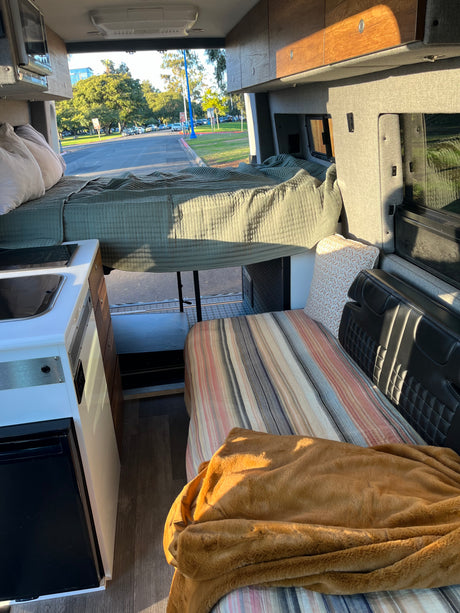 Sunlit RV rental interior featuring a raised bed, compact kitchen, and lower sleeping/seating area.