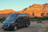 Dark gray Winnebago Solis RV rental on a dirt road, bathed in warm light from striking red rock mountains and green trees.